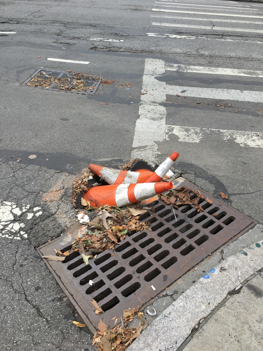 Three orange safetycones piled into a pothole next to a street drain. One is flattened.